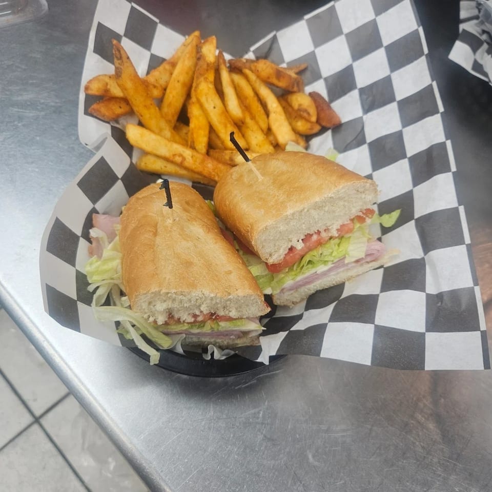 A sub sandwich with lettuce, tomato, and meat, served with a side of fries on checkered paper.