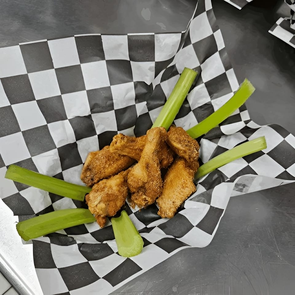 Crispy fried chicken wings with celery sticks in a black and white checkered paper-lined basket.