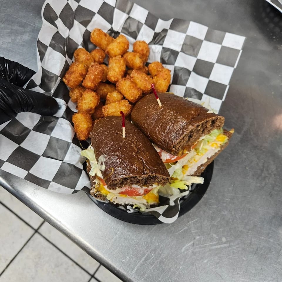 Two dark bread sandwiches with lettuce, tomato, and cheese, served with tater tots in a basket.