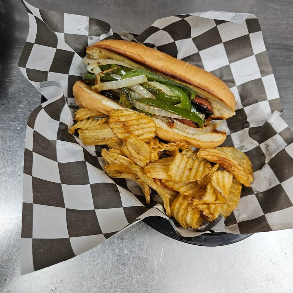 Hot dog with grilled onions and peppers, served with waffle-cut chips in a checkered paper-lined basket.