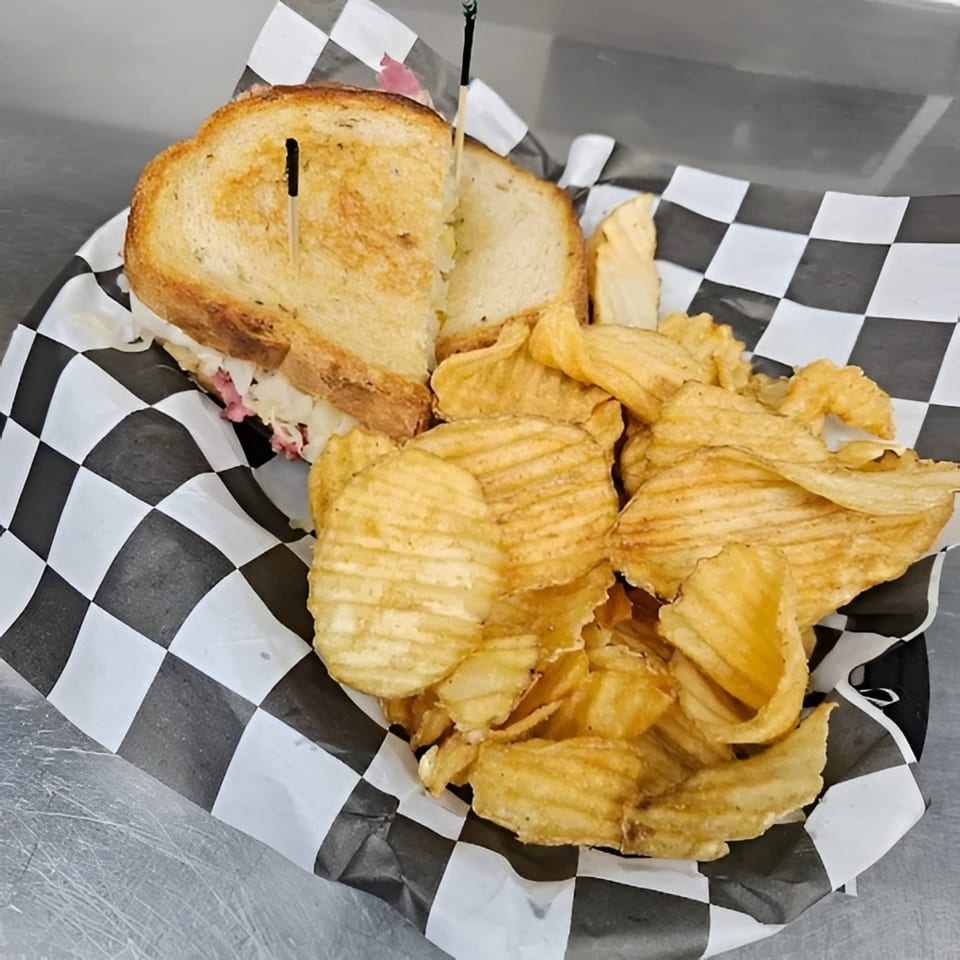 A grilled sandwich with chips in a checkered paper-lined basket on a metal surface.